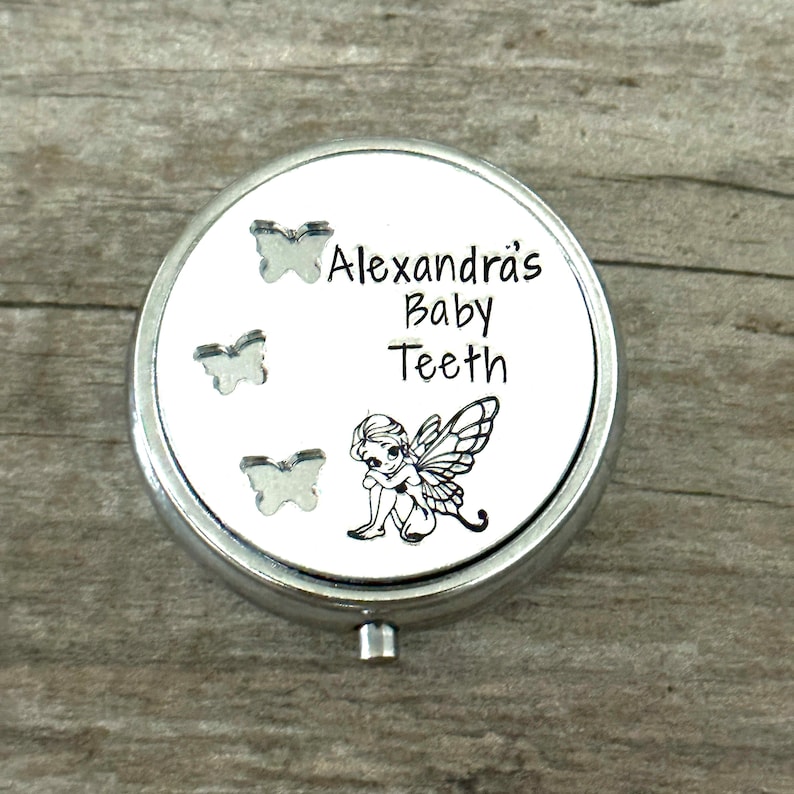 Puede incluir: Una caja redonda plateada para guardar dientes de beb&eacute;. La tapa presenta el texto "Alexandra's Baby Teeth" con tres recortes de mariposas y una ilustraci&oacute;n de hada. La caja est&aacute; sobre una superficie de madera.