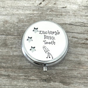 Puede incluir: Caja plateada para guardar dientes de leche con la inscripci&oacute;n "Zachary's Baby Teeth" y una ilustraci&oacute;n de un cohete. La tapa tiene tres recortes en forma de estrella. La caja redonda tiene un peque&ntilde;o asa plateada.