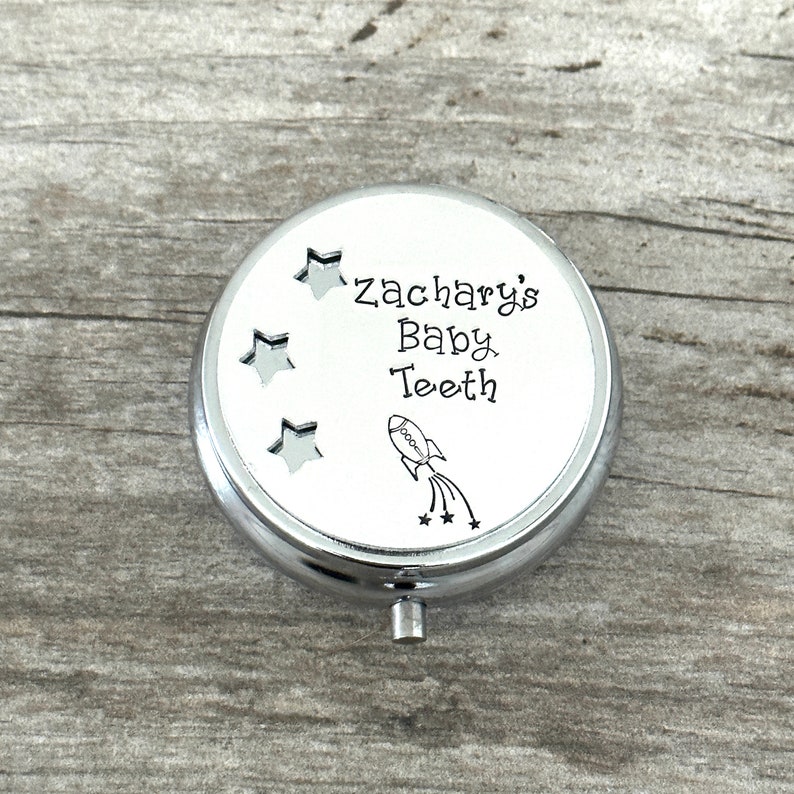 Puede incluir: Caja plateada para guardar dientes de leche con la inscripci&oacute;n "Zachary's Baby Teeth" y una ilustraci&oacute;n de un cohete. La tapa tiene tres recortes en forma de estrella. La caja redonda tiene un peque&ntilde;o asa plateada.