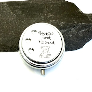 May include: A silver-colored, round keepsake box with the text "George's First Haircut" and a teddy bear illustration. Three heart-shaped cutouts are on the left side of the lid. The box has a small, silver handle.