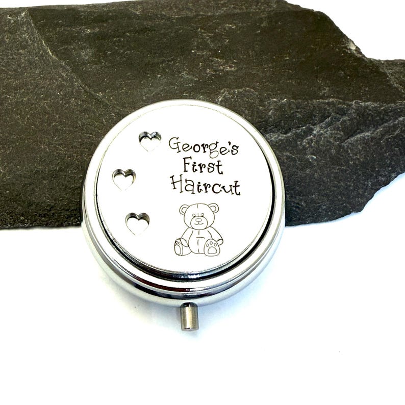 May include: A silver-colored, round keepsake box with the text "George's First Haircut" and a teddy bear illustration. Three heart-shaped cutouts are on the left side of the lid. The box has a small, silver handle.