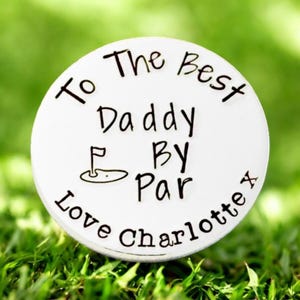 May include: A white golf ball marker with black text that reads "To The Best Daddy By Par Love Charlotte x". The marker features a golf course graphic and is set on a bed of green grass.