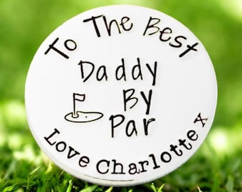 May include: A white golf ball marker with black text that reads "To The Best Daddy By Par Love Charlotte x". The marker features a golf course graphic and is set on a bed of green grass.