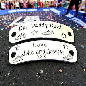 May include: Two curved silver-toned metal shoe tags with black engraved text and graphics. The top tag reads Run Daddy Run! with sneakers and stars. The bottom tag reads Love Jake and Joseph XXX with sneakers and stars. Each tag has two circular holes.