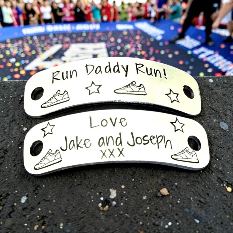 May include: Two curved silver-toned metal shoe tags with black engraved text and graphics. The top tag reads Run Daddy Run! with sneakers and stars. The bottom tag reads Love Jake and Joseph XXX with sneakers and stars. Each tag has two circular holes.