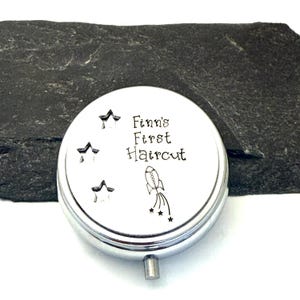 May include: A silver-colored, round keepsake box with the text "Finn's First Haircut" and star and rocket ship illustrations. The box has a small, silver-colored handle and is set against a dark gray background.