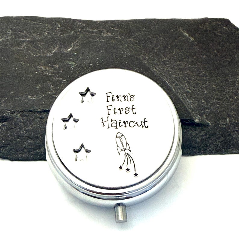May include: A silver-colored, round keepsake box with the text "Finn's First Haircut" and star and rocket ship illustrations. The box has a small, silver-colored handle and is set against a dark gray background.