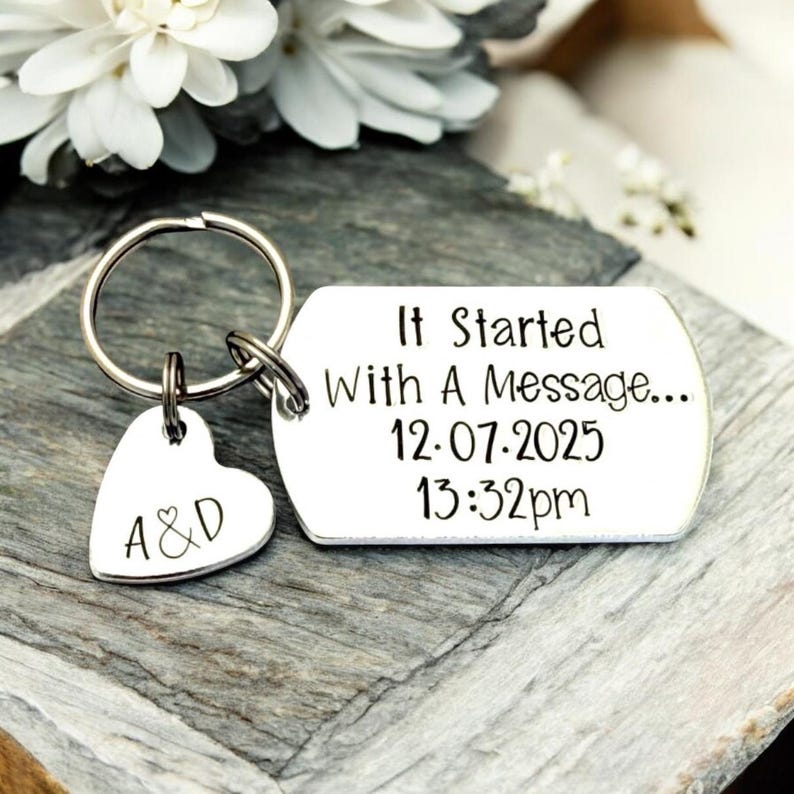 Puede incluir: Un llavero plateado con un dije en forma de coraz&oacute;n grabado con "A&D" y una etiqueta rectangular. La etiqueta dice "It Started With A Message... 12.07.2025 13:32pm". El llavero est&aacute; sobre una superficie gris texturizada.