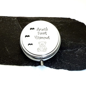 May include: A silver-colored, round keepsake box with the text "Amea's First Haircut" and a small teddy bear illustration. Three heart-shaped cutouts are above the text. The box is on a dark, textured surface.