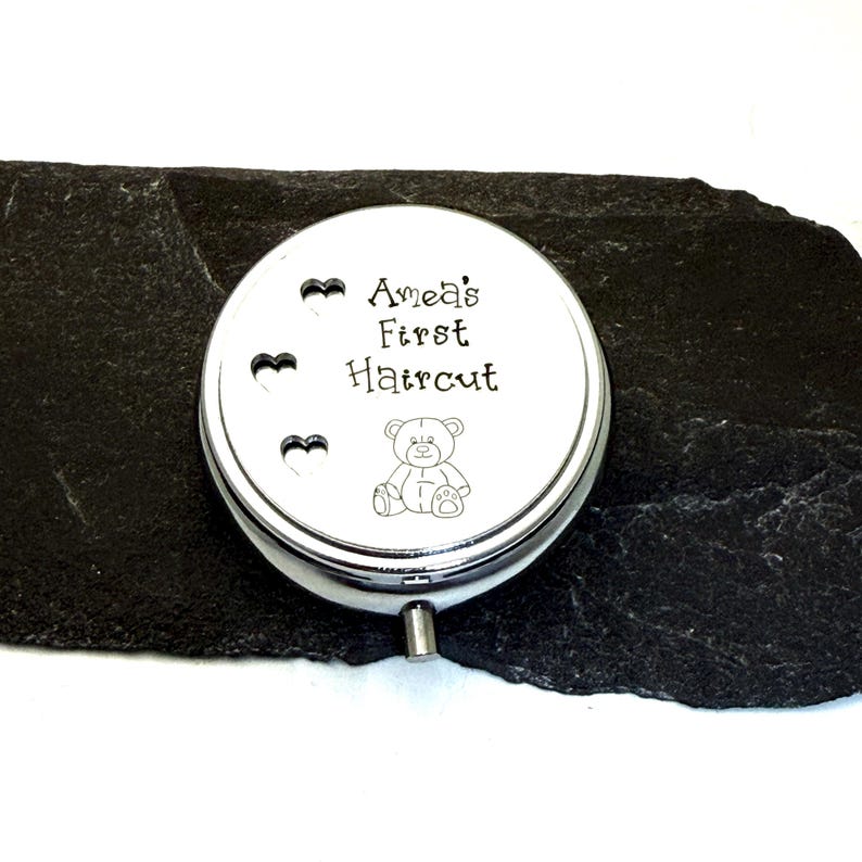 May include: A silver-colored, round keepsake box with the text "Amea's First Haircut" and a small teddy bear illustration. Three heart-shaped cutouts are above the text. The box is on a dark, textured surface.