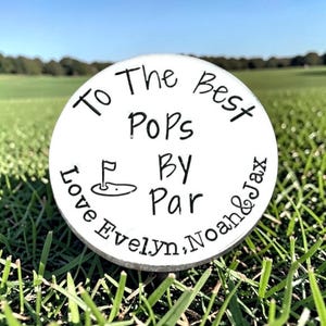 May include: A round white golf ball marker featuring black text that reads To The Best Pops By Par, with a small graphic of a golf flag on a green. The bottom of the marker includes the names Love Evelyn, Noah & Jax in black lettering.