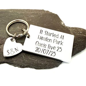 Puede incluir: Un llavero plateado con un dije en forma de coraz&oacute;n grabado con "S&N" y una etiqueta rectangular. La etiqueta dice "It Started At Heaton Park Oasis live 25 20/07/25" en texto negro. El llavero est&aacute; sobre una superficie de piedra gris oscuro.