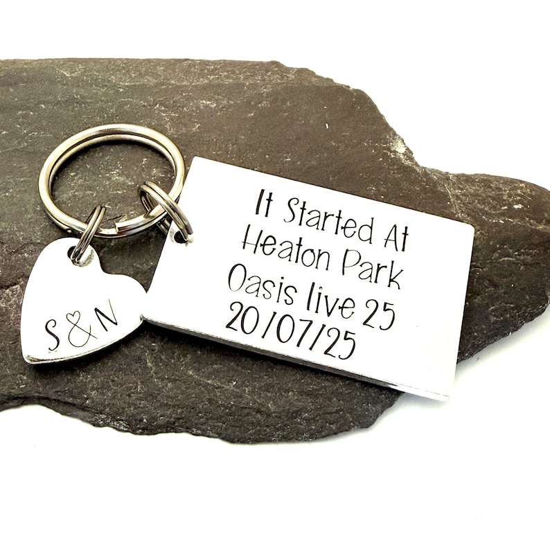 Puede incluir: Un llavero plateado con un dije en forma de coraz&oacute;n grabado con "S&N" y una etiqueta rectangular. La etiqueta dice "It Started At Heaton Park Oasis live 25 20/07/25" en texto negro. El llavero est&aacute; sobre una superficie de piedra gris oscuro.