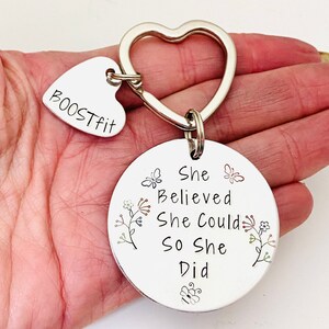 Pode incluir: Um porta-chaves de metal prateado com um anel em forma de cora&ccedil;&atilde;o, um pequeno pendente em forma de cora&ccedil;&atilde;o gravado com o texto BOOSTFIT e um pendente circular gravado com a frase She Believed She Could So She Did, decorado com pequenos desenhos de borboletas e flores.