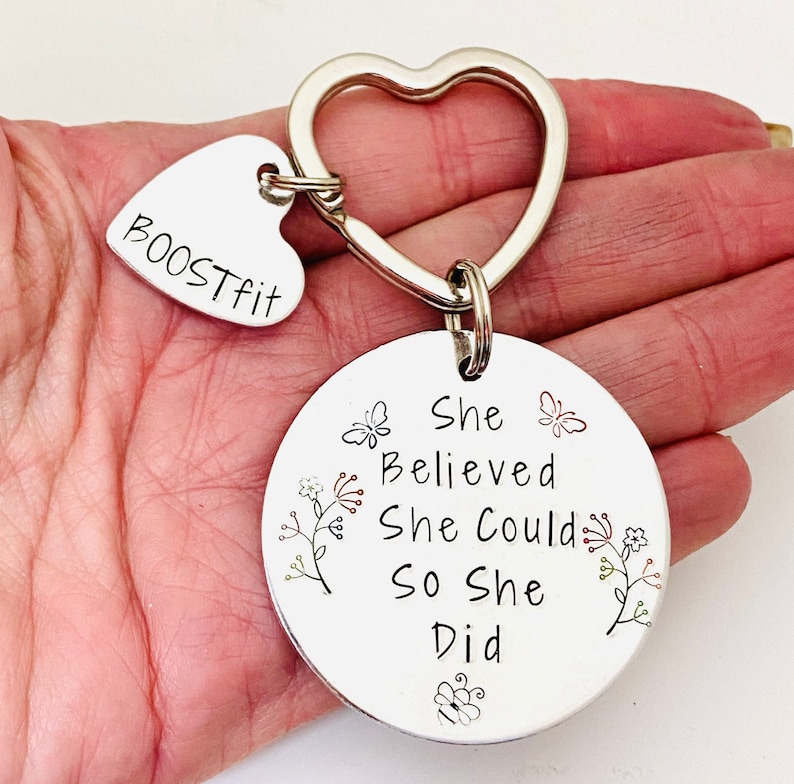 Pode incluir: Um porta-chaves de metal prateado com um anel em forma de cora&ccedil;&atilde;o, um pequeno pendente em forma de cora&ccedil;&atilde;o gravado com o texto BOOSTFIT e um pendente circular gravado com a frase She Believed She Could So She Did, decorado com pequenos desenhos de borboletas e flores.