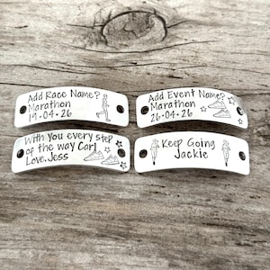 May include: Four silver-colored metal shoe tags with black text and illustrations. The tags include phrases like "Add Race Name?" and "Keep Going Jackie." Each tag has two holes for attaching to shoelaces. The background is a light brown wooden surface.