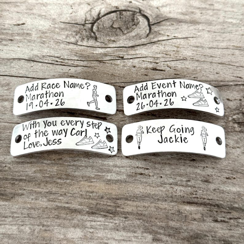 May include: Four silver-colored metal shoe tags with black text and illustrations. The tags include phrases like "Add Race Name?" and "Keep Going Jackie." Each tag has two holes for attaching to shoelaces. The background is a light brown wooden surface.