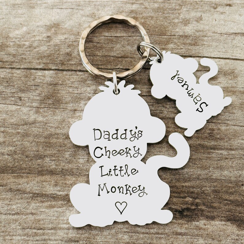 Puede incluir: Llavero de mono plateado con el texto "Daddy's Cheeky Little Monkey" y un coraz&oacute;n. Un llavero de mono m&aacute;s peque&ntilde;o con el texto "Yours" est&aacute; unido al llavero de mono m&aacute;s grande.