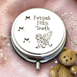 May include: A silver-colored baby tooth keepsake box with the text "Robyn's Baby Teeth" and a fairy illustration. The box has three star cutouts and a small handle. A teddy bear and sprigs of white flowers are in the background.