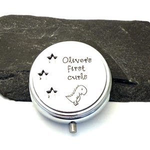 May include: A silver-colored, round keepsake box with the text "Oliver's first curls" and a small dinosaur illustration. The box has three star designs and a small, rectangular clasp. The background is a dark gray, textured surface.