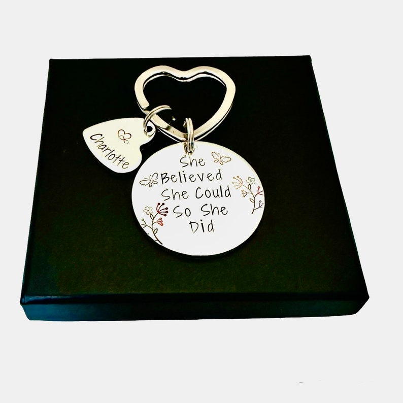 Pode incluir: Um porta-chaves de metal prateado com um anel em forma de cora&ccedil;&atilde;o, um pequeno pendente em forma de cora&ccedil;&atilde;o gravado com o nome Charlotte e um disco circular maior gravado com o texto She Believed She Could So She Did, decorado com motivos florais e borboletas.