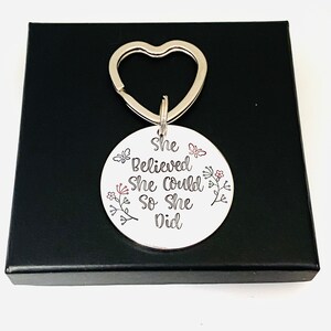 Pode incluir: Um porta-chaves circular de metal prateado com um anel em forma de cora&ccedil;&atilde;o. A superf&iacute;cie est&aacute; gravada com o texto preto "She Believed She Could So She Did" e decorada com pequenas ilustra&ccedil;&otilde;es de flores e borboletas em preto e vermelho.
