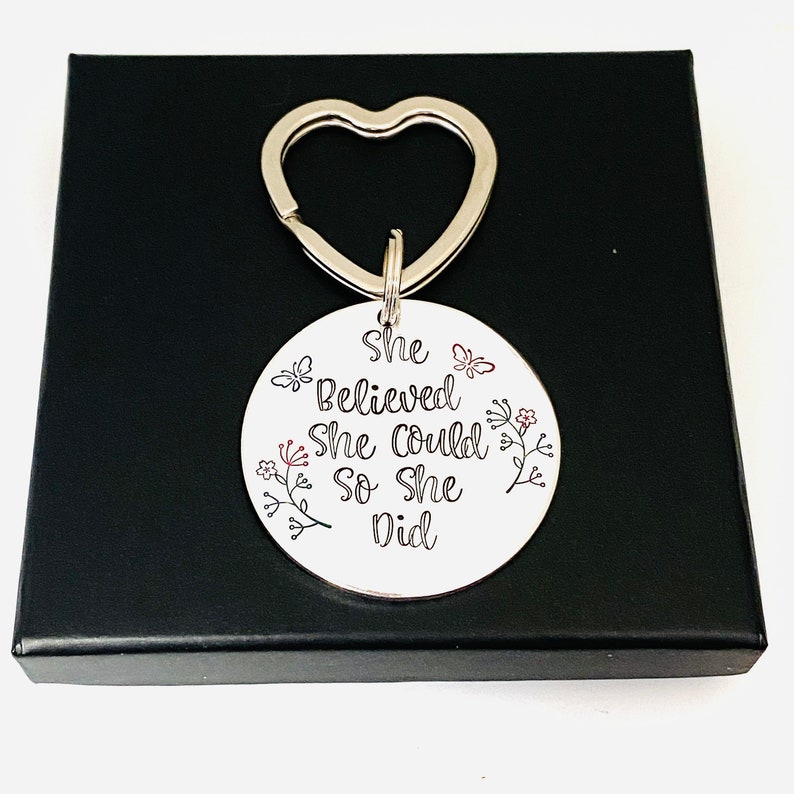 Pode incluir: Um porta-chaves circular de metal prateado com um anel em forma de cora&ccedil;&atilde;o. A superf&iacute;cie est&aacute; gravada com o texto preto "She Believed She Could So She Did" e decorada com pequenas ilustra&ccedil;&otilde;es de flores e borboletas em preto e vermelho.