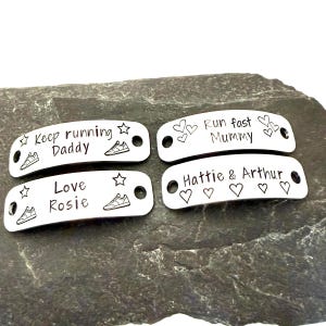 May include: Four silver-colored shoe tags with engraved text and designs. The tags feature phrases like "Keep running Daddy" and "Love Rosie", along with star and sneaker illustrations. The tags are on a dark gray stone surface.