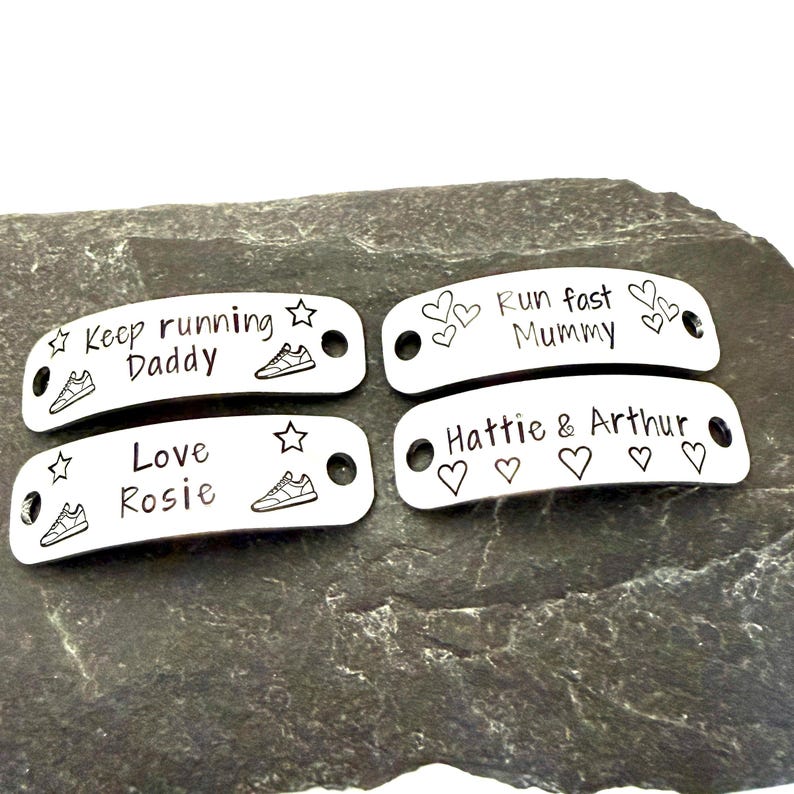 May include: Four silver-colored shoe tags with engraved text and designs. The tags feature phrases like "Keep running Daddy" and "Love Rosie", along with star and sneaker illustrations. The tags are on a dark gray stone surface.