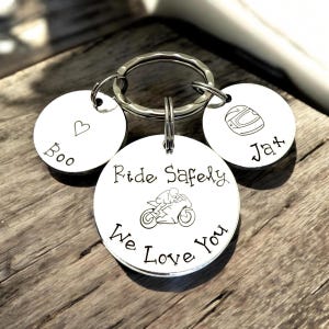 May include: A silver-toned metal keychain featuring three circular charms attached to a split ring. The largest center charm is engraved with a motorcycle graphic and the text Ride Safely We Love You. The smaller charms feature a heart with the name Boo and a motorcycle helmet with the name Jax.