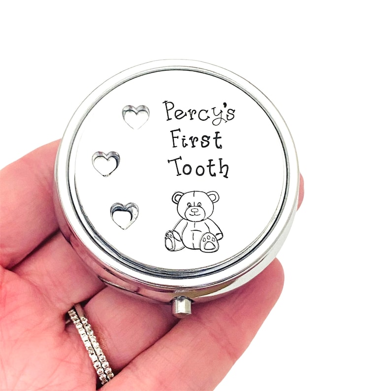 Puede incluir: Una caja de recuerdos redonda de color plateado con el texto "Percy's First Tooth" y una peque&ntilde;a ilustraci&oacute;n de oso de peluche. Tres recortes en forma de coraz&oacute;n est&aacute;n en el lado izquierdo de la tapa. La caja se sostiene en una mano.
