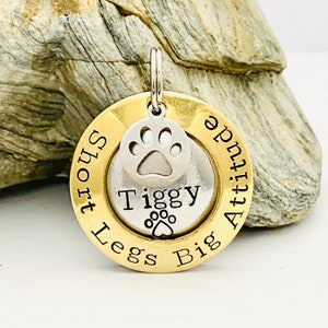 Pode incluir: Uma placa de identifica&ccedil;&atilde;o para c&atilde;es em ouro e prata com o texto "Short Legs Big Attitude" e o nome "Tiggy" gravado nela. Um amuleto de pata de prata est&aacute; preso &agrave; placa.