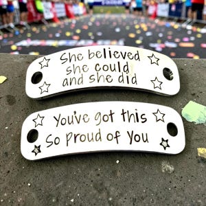 May include: Two curved silver-toned metal shoe tags with rounded ends and two circular holes. The top tag features the engraved text "She believed she could and she did" with four star icons. The bottom tag reads "You've got this So Proud of You" with four star icons.