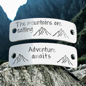 May include: Two silver metal tags with the phrases "The mountains are calling" and "Adventure awaits" in black lettering. Each tag features a mountain range graphic and a hole on each end.