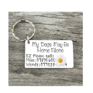 May include: A silver keychain with a white tag that reads "My Dogs May Be Home Alone ICE Please call: Mike: 07875 600: Wendy: 077025-4" with a white daisy graphic.