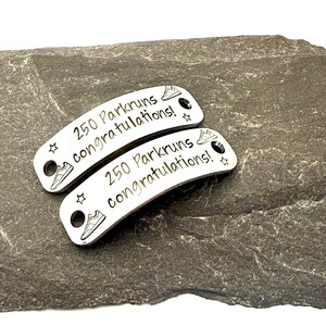 May include: Two silver Parkrun shoe tags with the text "250 Parkruns congratulations!" and a running shoe graphic. Each tag has a hole for attaching to a shoelace. The tags are on a dark gray stone surface.