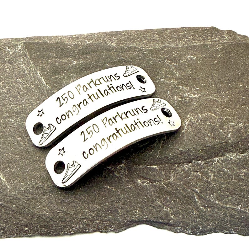 May include: Two silver Parkrun shoe tags with the text "250 Parkruns congratulations!" and a running shoe graphic. Each tag has a hole for attaching to a shoelace. The tags are on a dark gray stone surface.