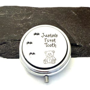 Puede incluir: Una caja redonda de recuerdo de color plateado con el texto "Justin's First Tooth" y una ilustraci&oacute;n de un oso de peluche. Tres recortes en forma de coraz&oacute;n est&aacute;n encima del texto. La caja est&aacute; sobre una superficie texturizada de color gris oscuro.