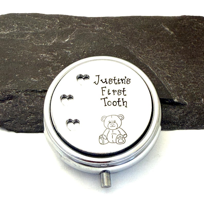 Puede incluir: Una caja redonda de recuerdo de color plateado con el texto "Justin's First Tooth" y una ilustraci&oacute;n de un oso de peluche. Tres recortes en forma de coraz&oacute;n est&aacute;n encima del texto. La caja est&aacute; sobre una superficie texturizada de color gris oscuro.