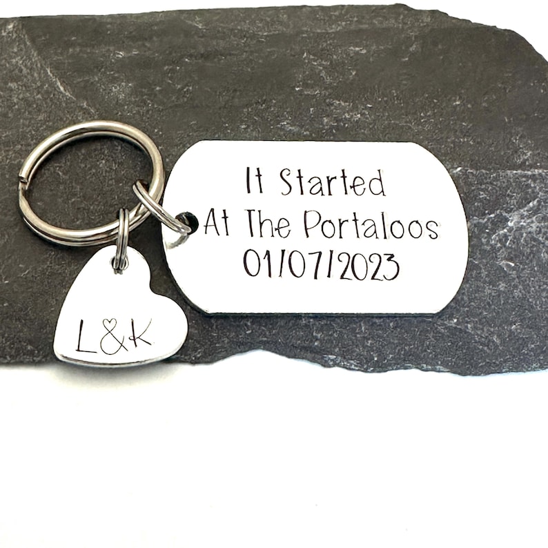 Puede incluir: Un llavero plateado con una etiqueta que dice "It Started At The Portaloos 01/07/2023". Un dije en forma de coraz&oacute;n con las iniciales "L&K" cuelga debajo. El llavero est&aacute; sobre una superficie de piedra gris oscura.