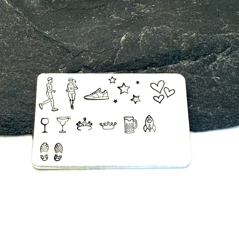 May include: A rectangular, silver-colored metal tag with various etched icons. The tag features images of a runner, a running shoe, stars, hearts, a wine glass, a cocktail glass, a crown, a beer mug, a rocket, and footprints.