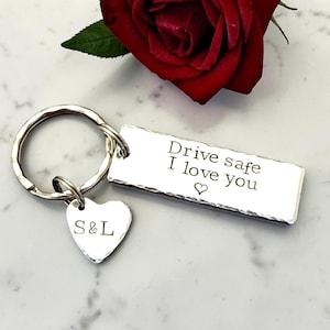May include: A silver keyring with a rectangular tag inscribed with "Drive safe I love you" and a heart symbol. A heart-shaped charm with "S&L" hangs below. A red rose is visible in the background.