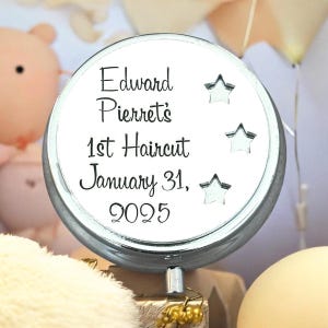 May include: A round, silver-toned metal keepsake box with a white lid featuring black text that reads Edward Pierret's 1st Haircut January 31, 2025. The lid is decorated with three small, silver-coloured star cutouts arranged vertically on the right side.