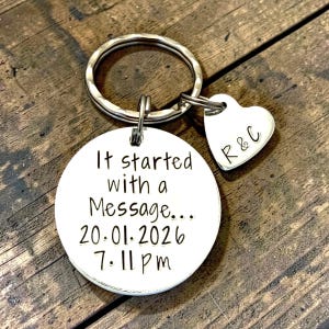 May include: A silver-toned metal keyring featuring a circular disc and a smaller heart-shaped charm. The circular disc is engraved with the text It started with a Message... 20.01.2026 7:11 pm. The heart charm is engraved with the letters R & C.
