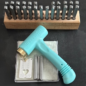 May include: A metal stamping kit with a light blue hammer, metal stamps, and a wooden holder. The kit includes metal blanks with the date 03/08/2013 and initials K and R. The hammer is labeled "ImpressArt".