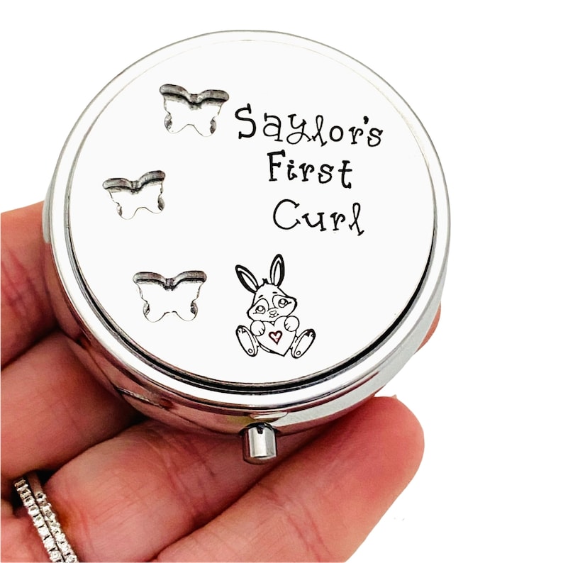 May include: A silver-colored, round keepsake box with the text "Saylor's First Curl." The lid features three butterfly cutouts and a cartoon bunny holding a heart. The box is designed to hold a baby's first curl.