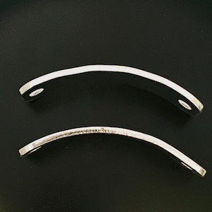 May include: Two curved, silver-colored metal pieces with a polished finish. Each piece has a hole at one end. The objects are set against a black background, highlighting their shape and reflective surface.