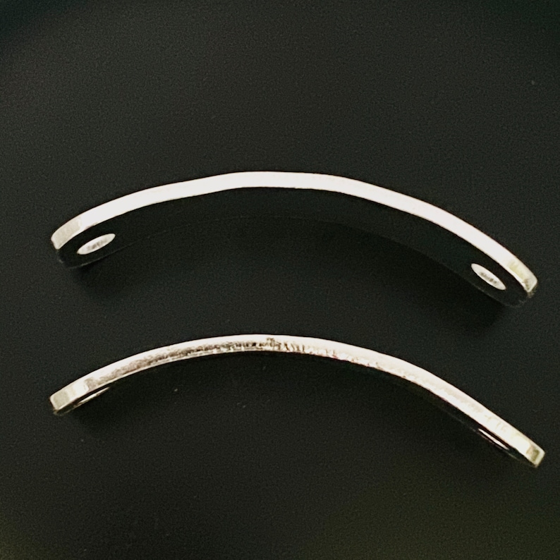 May include: Two curved, silver-colored metal pieces with a polished finish. Each piece has a hole at one end. The objects are set against a black background, highlighting their shape and reflective surface.