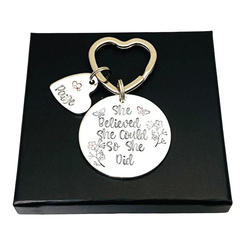 Pode incluir: Um porta-chaves de metal prateado com um anel em forma de cora&ccedil;&atilde;o, um pequeno pendente em forma de cora&ccedil;&atilde;o gravado com o nome Paige e um s&iacute;mbolo de infinito, e um disco circular gravado com o texto She Believed She Could So She Did e motivos florais.