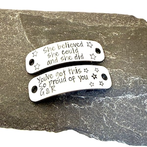 May include: Two silver-colored metal tags with inspirational quotes and star designs. The top tag reads "She believed she could and she did." The bottom tag says "You've got this. So proud of you G&R."